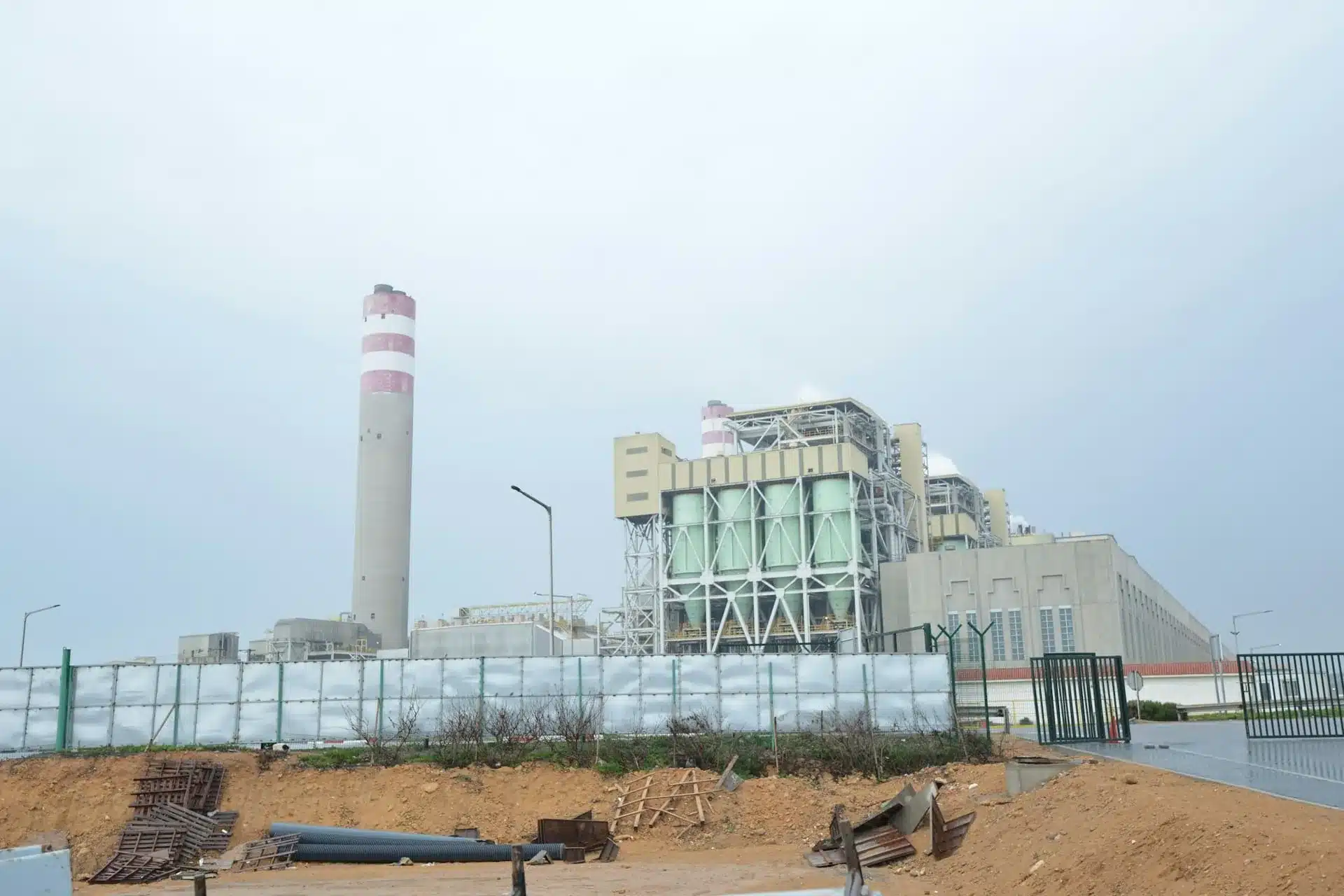 Le complexe industriel de Jorf Lasfar sous un ciel couvert : haute cheminée à bandes rouges et blanches, halles de la centrale et clôture de chantier au premier plan.